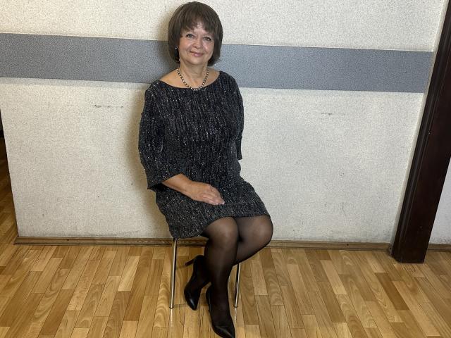 MaltaFora, middle-aged woman with short brown hair, black sparkly dress, black pantyhose, black heels, sitting on metal chair, wooden floor, smiling, necklace, hands on lap.
