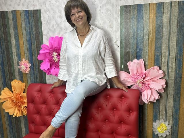 MaltaFora, middle-aged woman with short brown hair, white button-up shirt, light blue jeans, necklace, smiling, sitting on red tufted couch, large pink and orange flowers on either side.