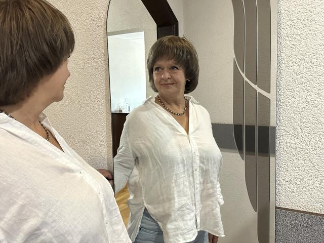 MaltaFora with short brown hair, white button-up shirt, necklace, standing in front of mirror, looking at reflection, showing her big boobs, and a subtle smile.