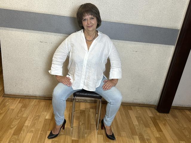 MaltaFora, short brown hair, white button-up shirt, light blue jeans, black high heels, sitting on metal chair, hands on knees, wooden floor, smiling.