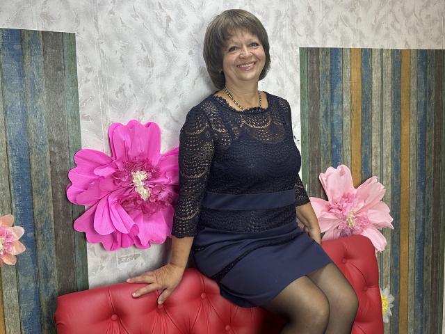 MaltaFora, middle-aged woman with short brown hair, wearing a navy lace dress and black pantyhose, sitting on a red tufted couch, smiling, with pink and pinkish flowers.