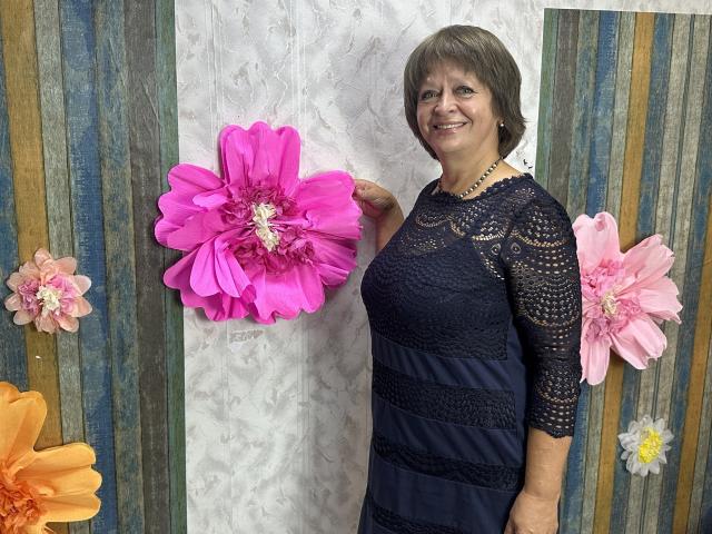 MaltaFora with short brown hair, smiling, wearing a dark lace and navy dress, holding a large pink flower. Pink and orange flowers on the left and right.