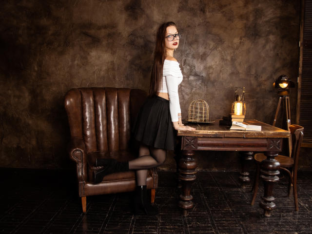 BethCutie con cabello largo y castaño, gafas, top corto blanco, falda negra, medias negras, tacones negros, arrodillada sobre una silla de cuero marrón, al lado de una mesa de madera con una linterna y libros..