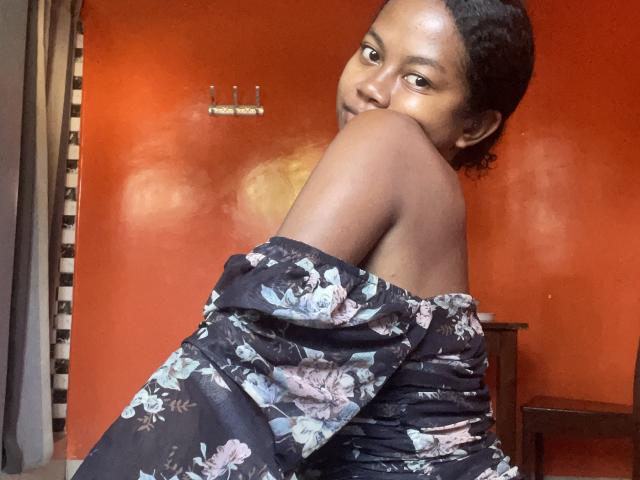 Sabaniah with dark skin, curly hair, floral off-shoulder top, looking over shoulder, brown eyes, small breasts, orange wall, wooden chair, metal hook in background.