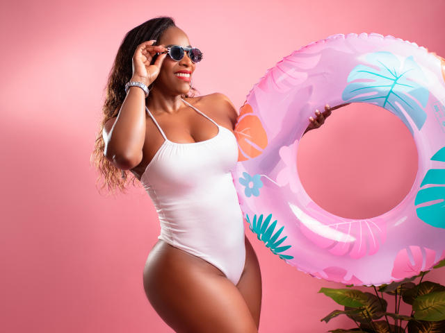 IsabellaKaned with big ass, white one-piece, holding pink inflatable with tropical patterns, wearing sunglasses, smiling, dark brown skin, wavy hair, pink background, green plant bottom right.