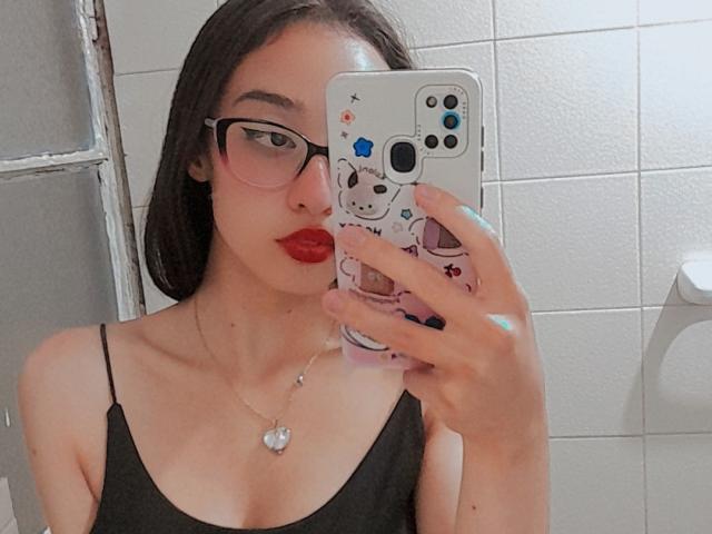 CamiVargas com batom vermelho, regata preta, óculos e colar, segurando um celular com capa de panda, tirando uma selfie no espelho, mostrando seios pequenos, com um fundo de parede branco com azulejos..