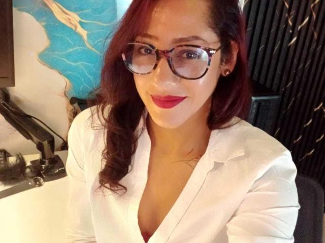 SamanthaCorine with red hair, glasses, and white button-up shirt, showing her tits in the low V-neck. She's smiling, wearing red lipstick, and has small gold earrings.