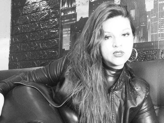 SkylarDesire with long, wavy hair, wearing a leather jacket, sitting in a black leather chair, looking slightly to the side with a subtle, confident expression. Black-and-white photo.