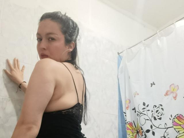 SylvieColette, con cabello negro largo, vestida con una camiseta de encaje negro, mirando por encima del hombro, tocando una pared de azulejos blancos, mostrando su espalda y trasero. Escena en un baño..