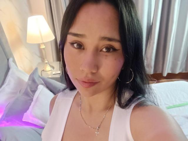 Karollteyes, avec ses longs cheveux noirs, portant un haut blanc, des boucles d'oreilles cerceaux et un collier avec une croix, a de petites seins, de l'acné rosée sur sa joue, et une expression mignonne, légèrement nerveuse..