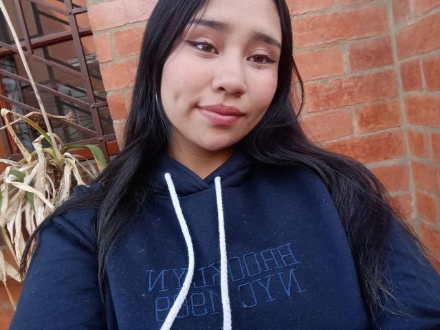 Karollteyes with long black hair, wearing a navy hoodie, has a subtle smile, and her brown eyes are slightly squinted. Her medium-sized, soft-looking tits are barely visible.