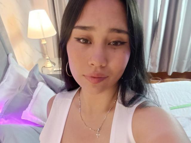 Karollteyes with long black hair, wearing a white tank top, hoop earrings, and a cross necklace, has small tits and a cute face, standing in a bedroom.