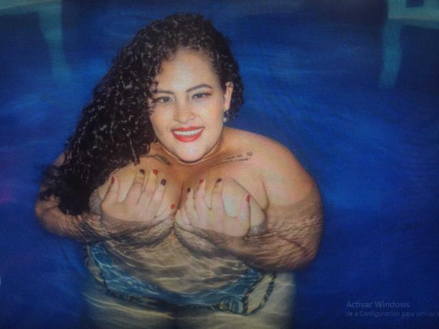 LucyGabry with curly black hair, red lipstick, and red nail polish, covering her large tits with hands in a blue swimming pool, smiling at the camera.