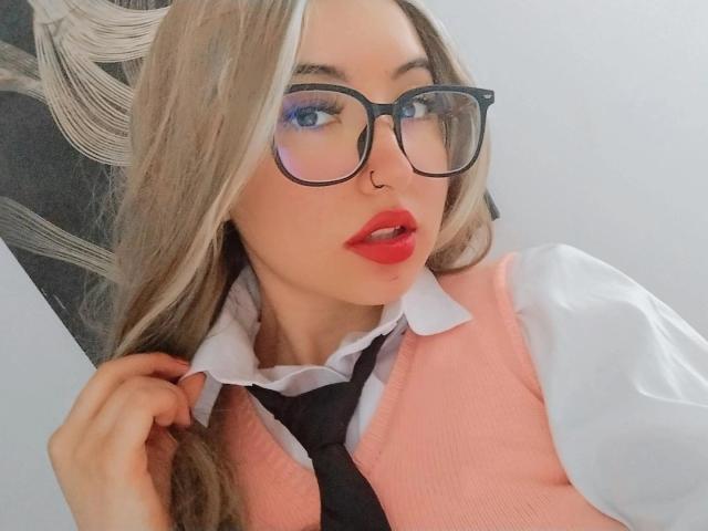 SusanMonroy with glasses, red lipstick, and nose ring, wearing a pink vest over a white shirt and black tie, touching her hair. Blonde hair, blue eyes, sexy.