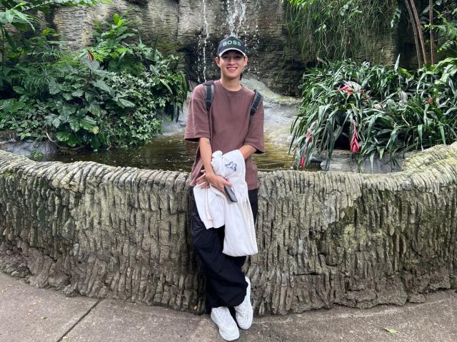 HaroldX stands against a rough stone wall, wearing a brown shirt, black pants, white sneakers, green cap, backpack, holding a white jacket, with a small waterfall behind.
