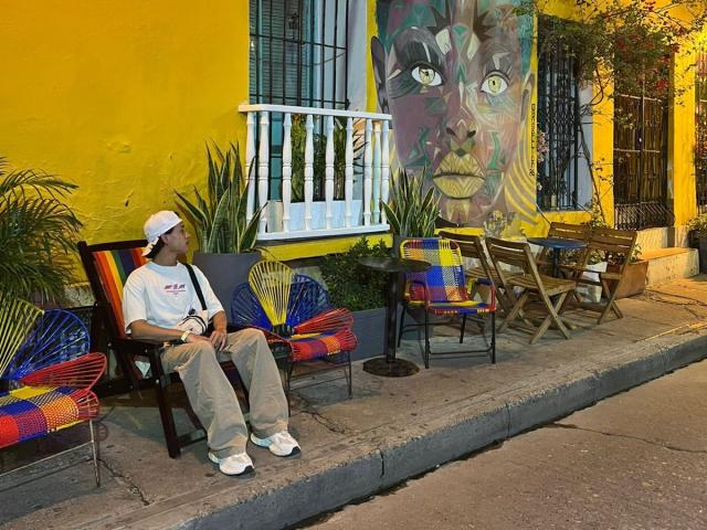 HaroldX, portant une casquette blanche, est assis sur une chaise colorée, un appareil photo à la main, devant un mur jaune vif orné d'une grande peinture abstraite représentant un visage..