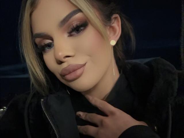 IvyRaven has blonde and black hair, thick eyebrows, long eyelashes, pink lipstick, and is wearing a black jacket with a zipper. She's touching her neck with a hand.