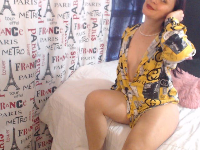 Wendy Schulz, vestindo um robe amarelo com padrão, decote em V profundo, mostrando os seios, sentada na cama, pernas afastadas, travesseiro rosa, cabelo escuro, batom vermelho, colar, cortina com Paris, Torre Eiffel e Fréch..