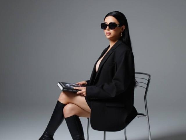 EvaPapaya with black sunglasses, long straight black hair, black blazer, no shirt, black knee socks, and black boots, sitting on a clear chair, holding a book.
