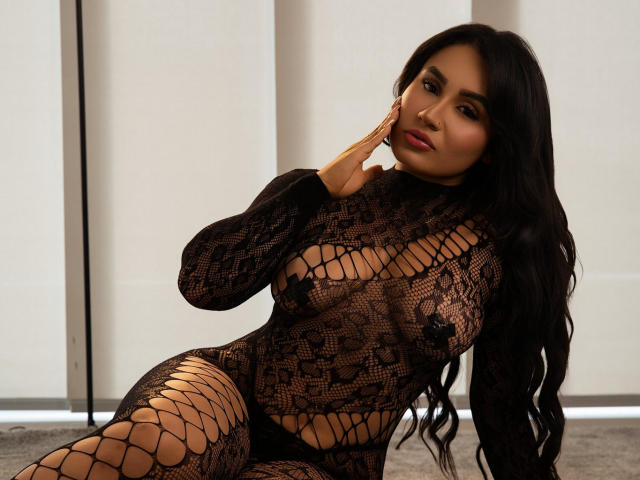 AshlyFerria with long black hair, wearing a black lace fishnet bodysuit, exposing her tits and pussy through the pattern, sitting on a carpet, hand on her cheek.