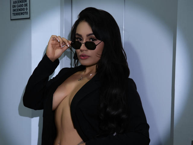 AshlyFerria with long black hair, wearing a black blazer open to show her tits, holding round sunglasses, looking at the camera with a slight smile.