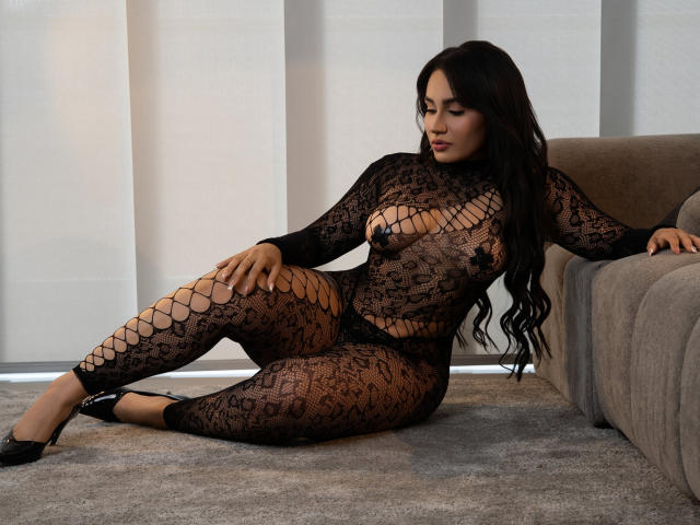 AshlyFerria with long black hair, wearing a see-through lace bodysuit, exposing her tits and pussy, sitting on a carpet, leaning on a gray couch, in black heels.