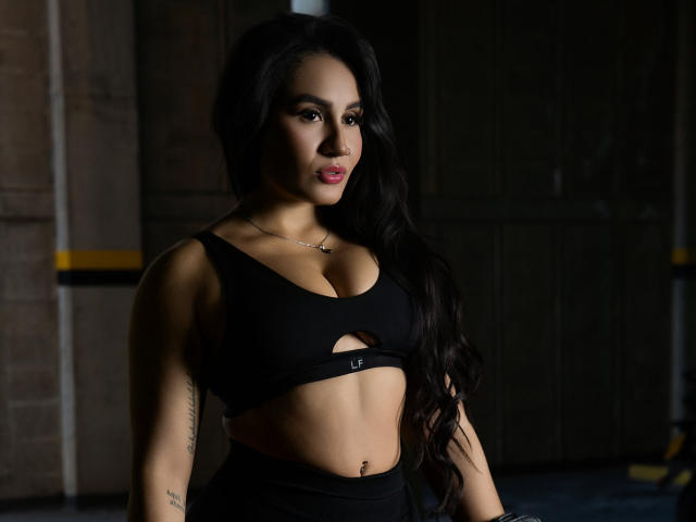 AshlyFerria with long black hair, wearing a black sports bra showing her tits and a small cutout, has tattoos on her arms, and a belly button piercing.