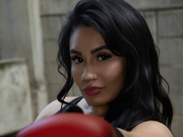 AshlyFerria has dark, wavy hair, brown eyes, and red lipstick, wearing a nose ring, looking serious with a red boxing glove in the foreground.