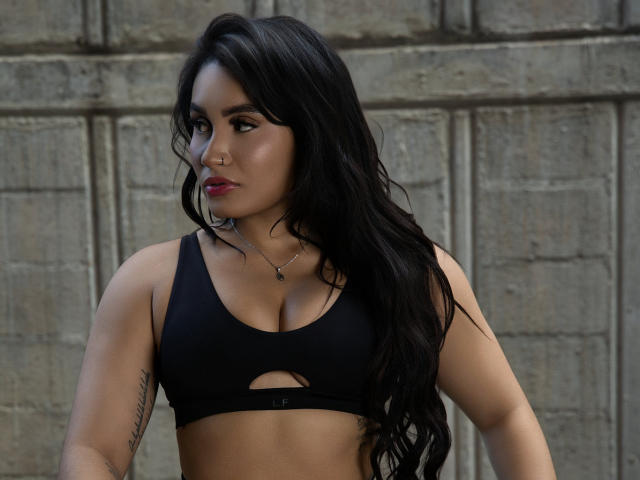 AshlyFerria with long black hair, tan skin, and red lipstick, wearing a black sports bra, showing her tits and a tattoo on her right arm, looking to the side.