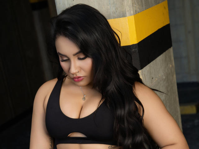 AshlyFerria with long black hair, downcast eyes, black sports bra, showing her tits, nose ring, and necklace, leaning against a yellow and black striped column.