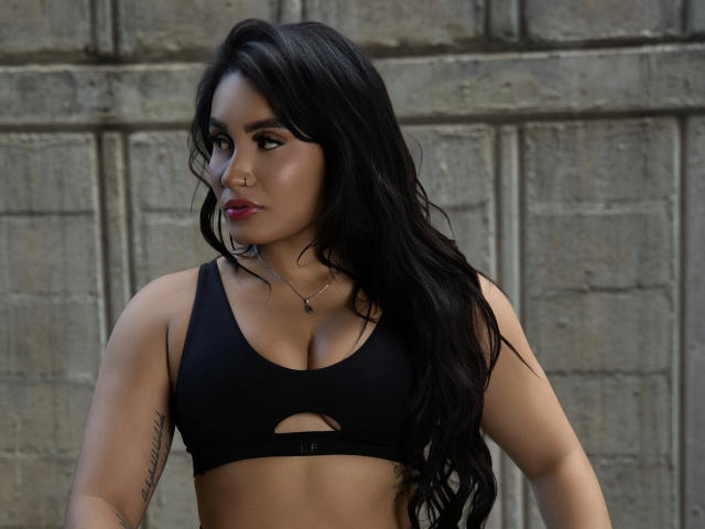 AshlyFerria with long black hair, wearing a black sports bra, showing her tits, nose ring, and tattoo on her right arm, looking to the side with red lipstick.