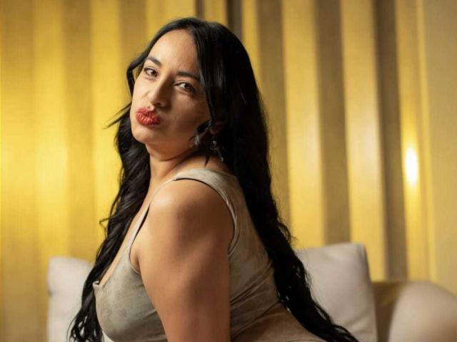 ChanelMilf with long black hair, red lipstick, and a beige top, looking over her shoulder, showing her ass. Soft yellow lighting in the background.