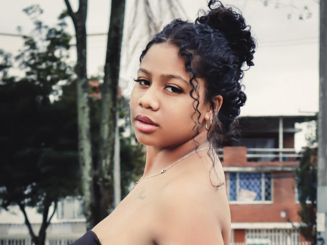 SofiFoxy69 with curly black hair in a bun, wearing a strapless black top, showing a small tattoo on her shoulder, looking over her shoulder with a serious expression.