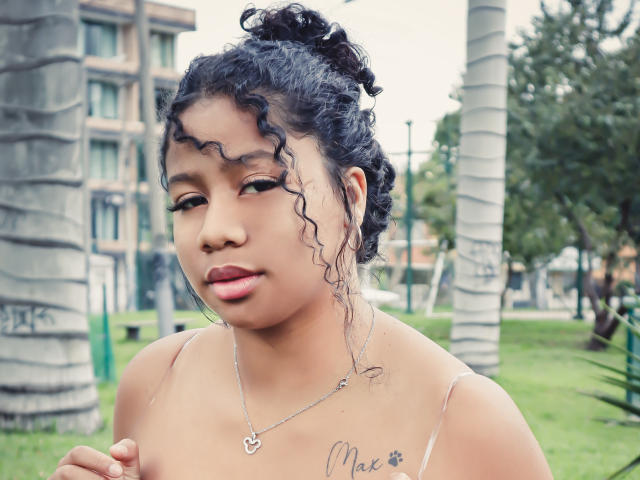 SofiFoxy69 with curly black hair in a bun, brown skin, red lipstick, and a necklace, showing her small tits and tattoo under her collarbone, in an outdoor setting.