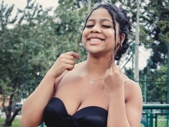 SofiFoxy69 with dark hair in a bun, smiling, wearing a black strapless top showing her tits, heart necklace, holding hair, green outdoor background, sunny day.