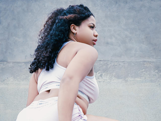 SofiFoxy69 with curly black hair, wearing a white sports bra and pink Calvin Klein shorts, showing her big ass and small tits, looking to the side with a pout.