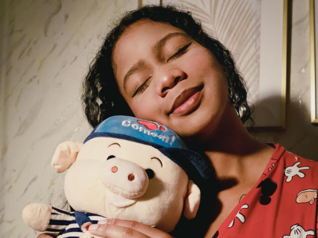 SofiFoxy69 with curly black hair, eyes closed, smiling, holding a white stuffed bear wearing a blue hat, in a red shirt with cartoon prints, sunlight on her face.