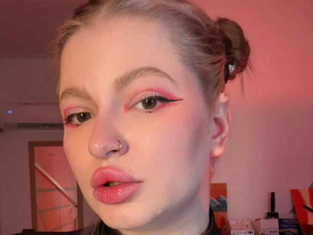 MiaKamp has pink eyeliner, a nose ring, and pink lips. She's looking at the camera with her blonde hair in two buns. Her face is slightly flushed.