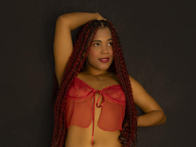 KatiEvans with long red braids, wearing a red lace crop top, exposing her tits, hand on head, dark background, smiling with red lipstick.