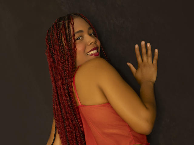 KatiEvans with red braided hair, smiling at the camera, wearing a red sheer top, hand on dark wall, showing her ass, tits, and cute face.