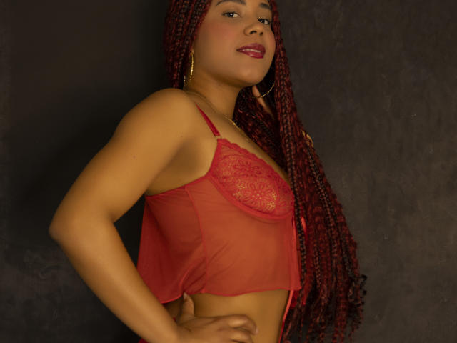 KatiEvans with red braided hair, wearing a red lace bralette, hands on hip, smiling, dark background, gold hoop earrings, medium brown skin, red lipstick.