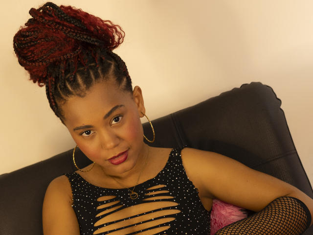 KatiEvans with red and black braided hair, gold hoop earrings, and a black cut-out dress, sitting on a black leather couch, wearing fishnet arm sleeves.
