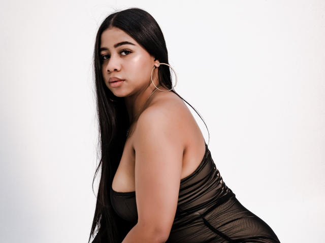 SofiFoxy69 with long black hair, wearing a sheer black dress, showing her tits, large hoop earrings, and a serious expression, against a plain white background.