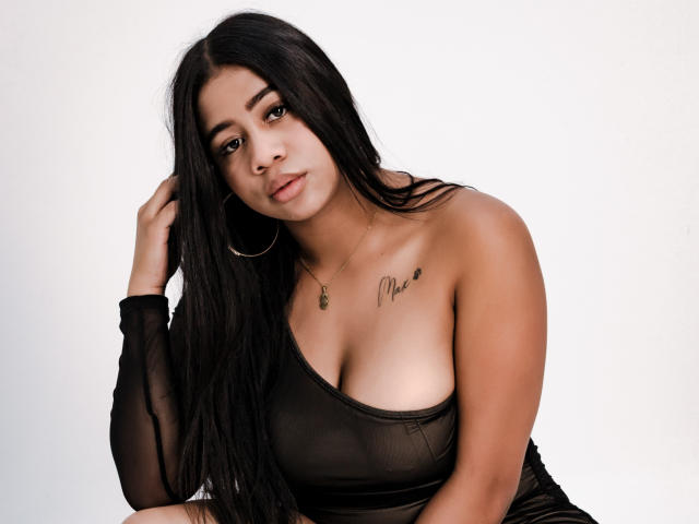 SofiFoxy69 with long black hair, hoop earrings, and a black sheer top showing her tits, a small tattoo near her right breast, and a necklace. She's looking at the camera.