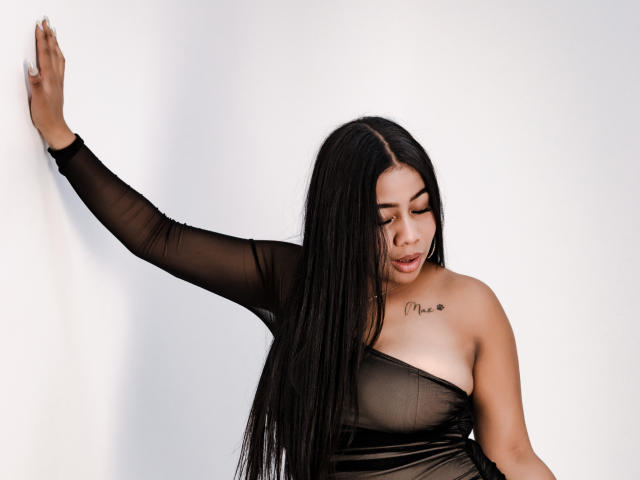 SofiFoxy69 with long black hair, wearing a sheer black dress, has her tits slightly visible, arm raised, looking down, tattoo on chest saying "Chloe".