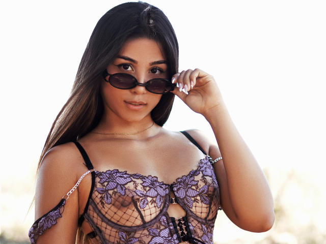 AnaWillsson with long dark hair, wearing purple lace bra with cut-out, black straps, holding sunglasses, smiling, showing small breasts, tan skin, brown eyes, outdoors, daytime.