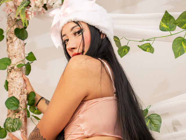 AnnieSkov met lang zwart haar, een roze satijnen beha, een witte pluizige hoed, rode lippenstift en groene blad-tattoos, kijkt over haar schouder en houdt een boom met groene bladeren vast..
