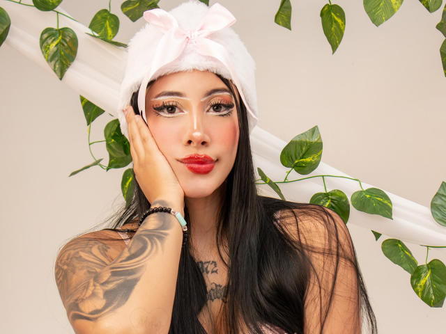 AnnieSkov, con lunghi capelli neri, indossa un cappello bianco e soffice con un fiocco rosa, rossetto rosso, un braccio tatuato e, sullo sfondo, rampicanti verdi e fogliosi. La sua mano tocca la guancia..