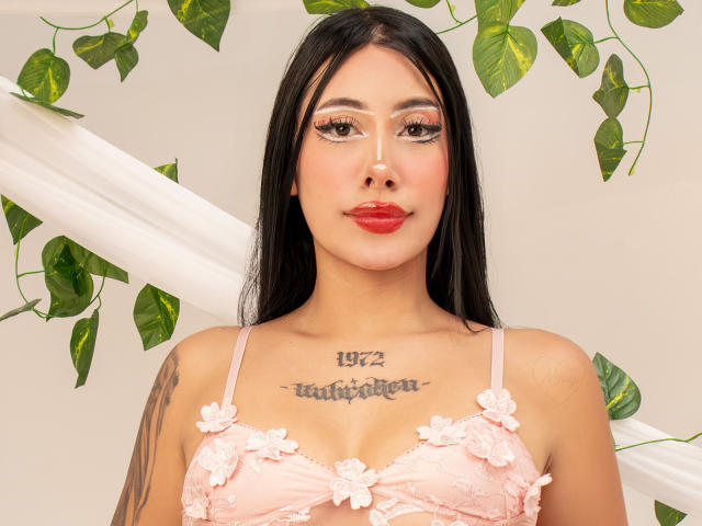 AnnieSkov with long black hair, pink floral bra, red lipstick, tattoos, and a nose ring, showing off her tits and a tattoo above her boobs.