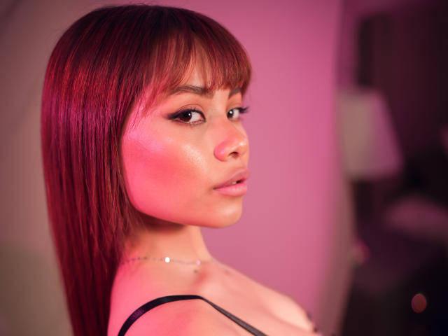 VannesaSaenz has red hair, bangs, and a shiny face with pink lighting, wearing a black strap top, looking over her shoulder with a slight pout.